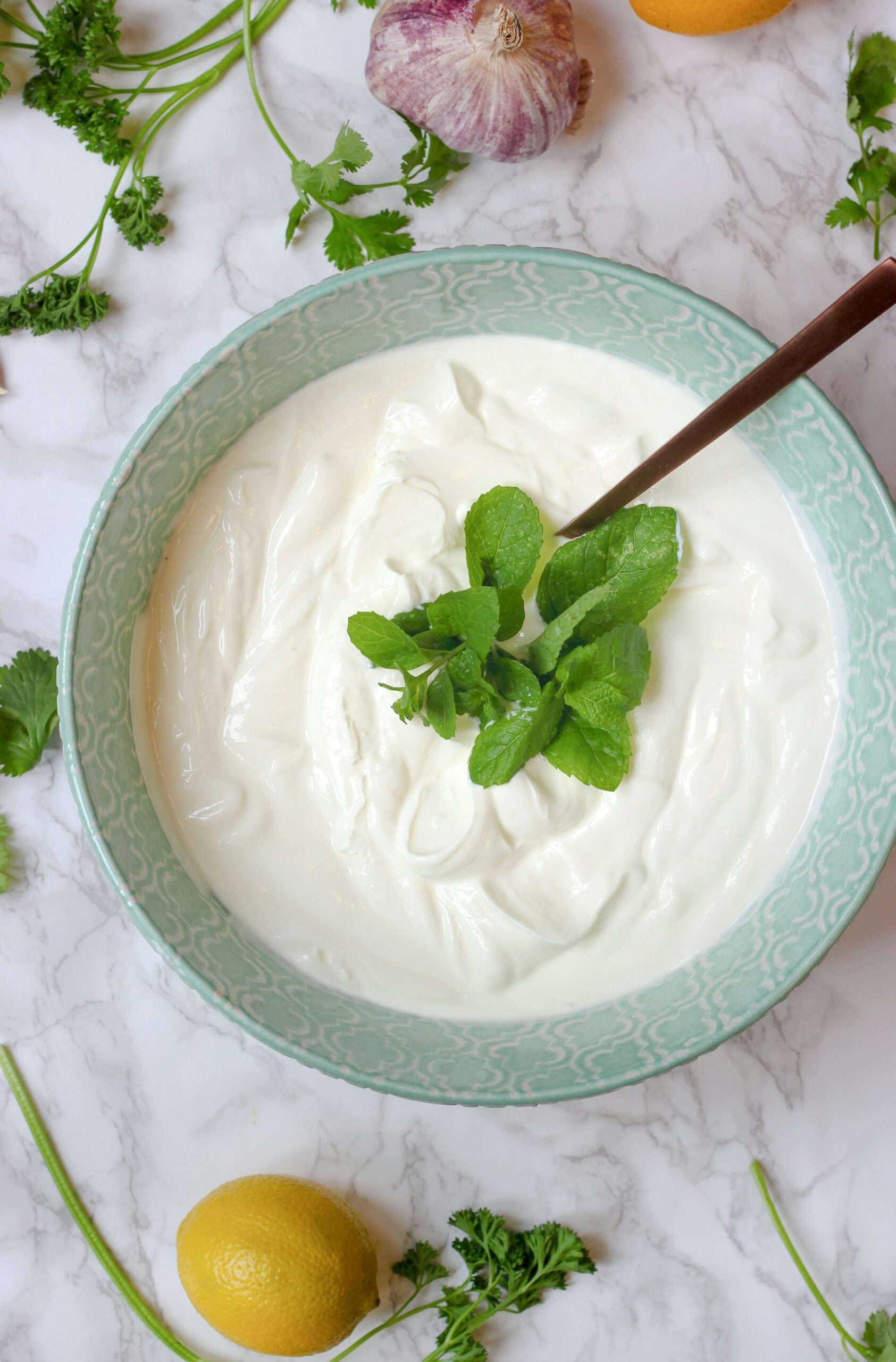 A pale green patterned bowl of thick white yogurt topped with fresh mint, a spoon resting in it, on white marble with a lemon, garlic and parsley