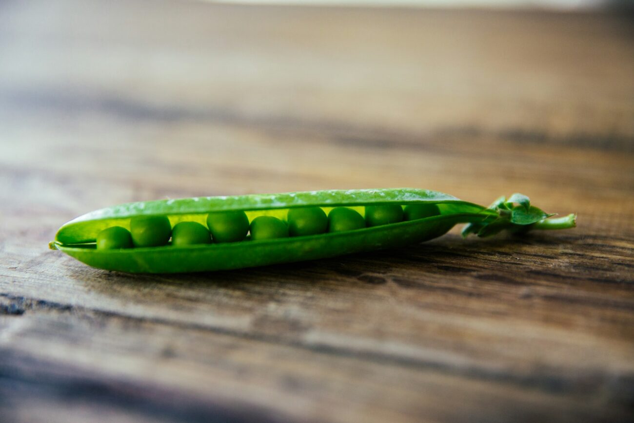 One pea pod split open on a wooden table, the row of green peas showing inside