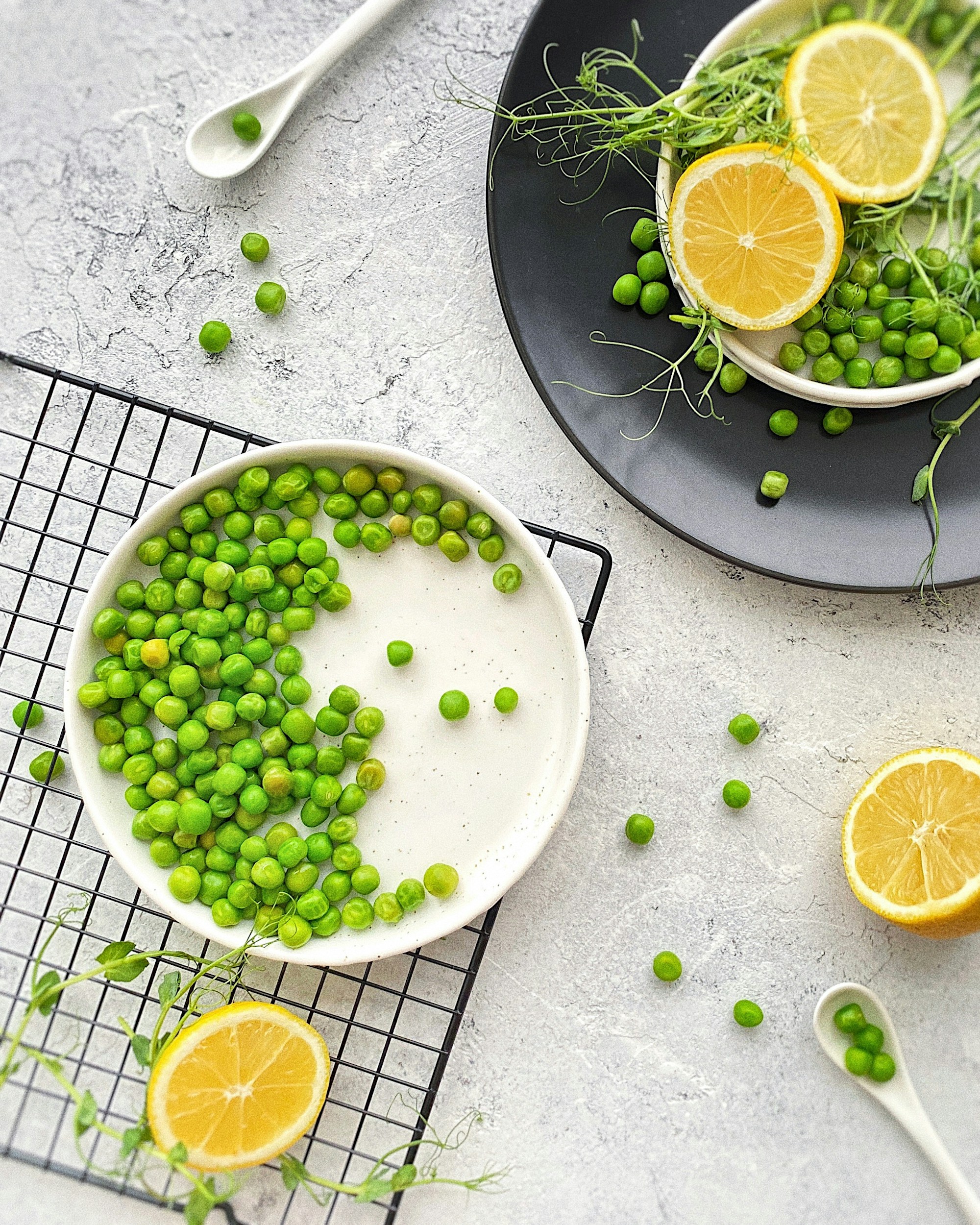 Shelled green peas in a white bowl on a wire rack, with pea shoots and lemon halves around it
