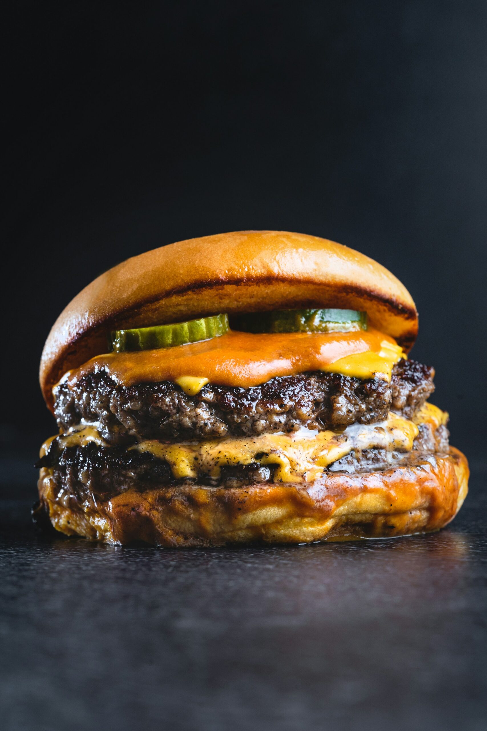 A double cheeseburger on a dark surface, two seared beef patties under melted orange cheese and pickle slices, in a glossy bun
